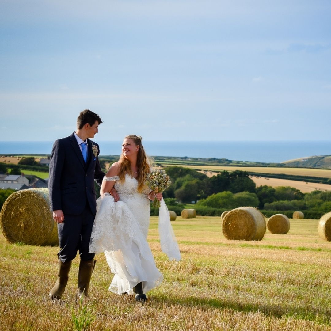 Bride & groom walking in a field with straw bales with sea in the distance